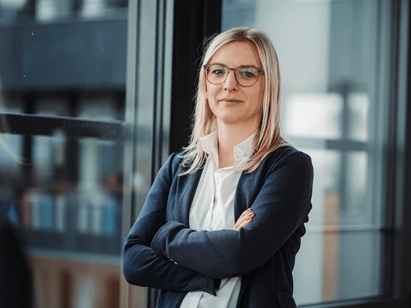 blonde Frau mit Blazer steht vor Fensterfront mit verschränkten Armen