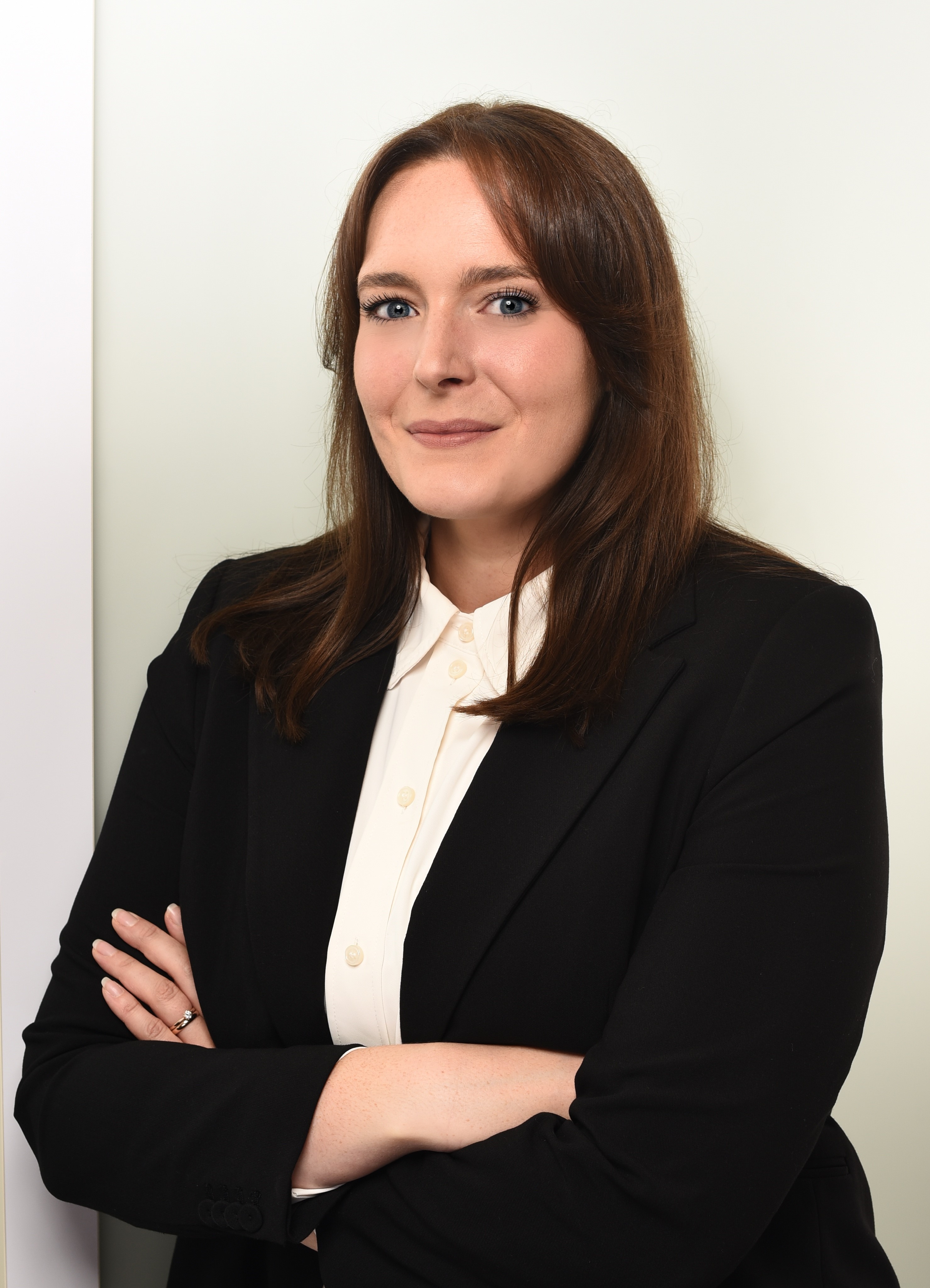 Eine beruflich gekleidete Frau mit langen, braunen Haaren steht mit verschränkten Armen und lächelt freundlich in die Kamera. Sie trägt ein schwarzes Blazer und ein weißes Hemd, während sich im Hintergrund eine helle Wand befindet.